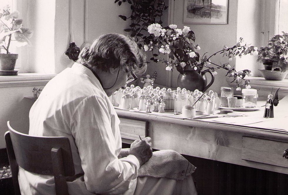 Schwarz-Weiß-Aufnahme. Rückansicht von Olly während der Arbeit. Auf dem Tisch vor ihr stehen Blumen und zahlreiche unbemalte Figuren.