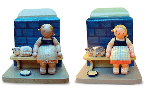 Figurenensemble „Aschenbrödel“. Aschenbrödel sitzt auf einer Holzbank. Sie lehnt mit dem Rücken an einem blauen Kachelofen. Sie trägt eine Schürze mit Karomuster und einen roten Rock. Neben ihr schläft eine graumelierte Katze. Am Boden steht ein