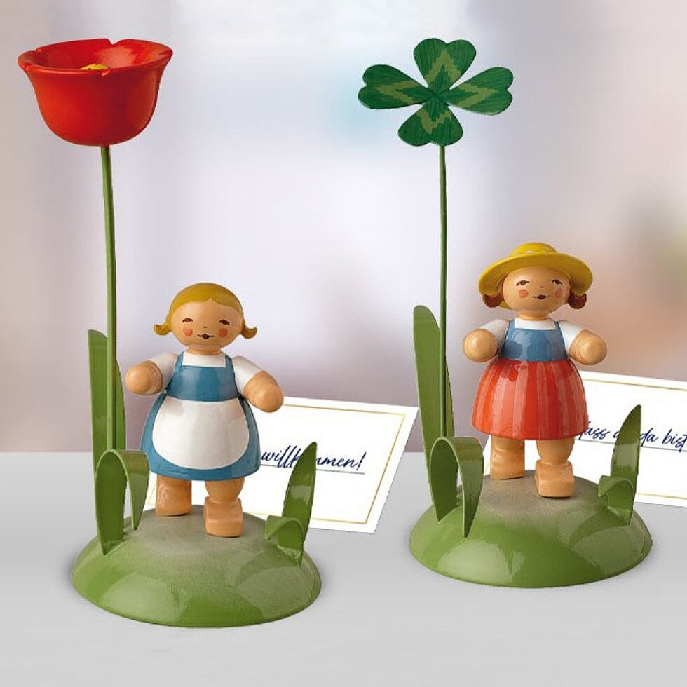 Zwei handgefertigte Tischkartenhalter mit kleinen Holzfiguren, einer mit roter Blume, der andere mit grünem Kleeblatt, beide auf grünen runden Sockeln