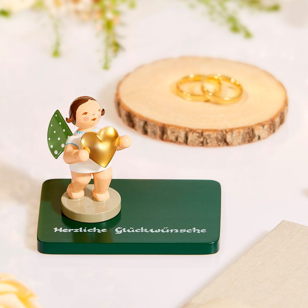 Holzengel mit grünen Flügeln von Wendt & Kühn hält goldenes Herz auf grünem Sockel mit der Aufschrift 'Herzliche Glückwünsche', im Hintergrund zwei goldene Ringe auf Holzscheibe