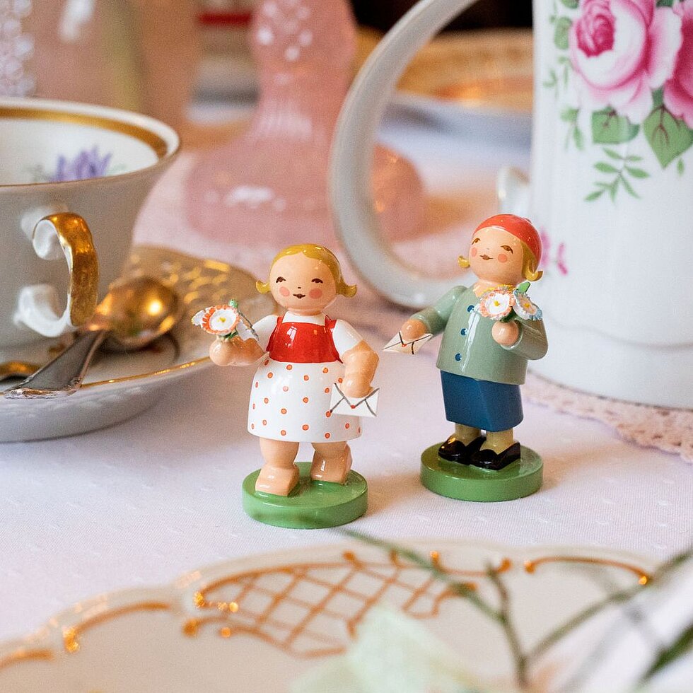 Zwei handgefertigte Holzfiguren eines Mädchens und eines Jungen mit Blumen und Briefen auf einem gedeckten Tisch