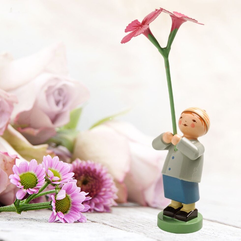 (c) Figur: Wendt & Kühn KG, Hintergrund: gudrun (Adobe Stock) Figur eines Jungen mit blauem Rock und grauer Jacke, der eine große pinke Blume hält, vor verschwommenen rosa und lila Blumen im Hintergrund