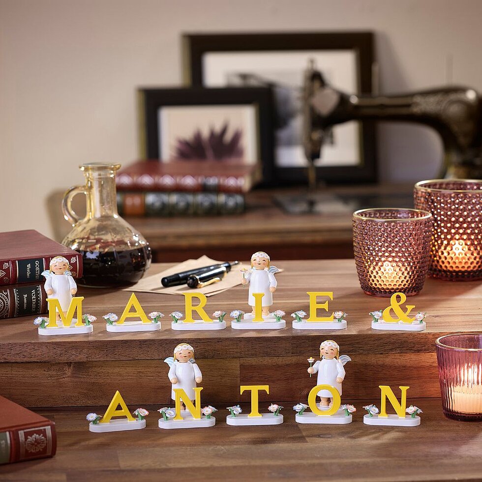 Holzfiguren von Engeln mit gelben Buchstaben, die die Namen 'MARIE & ANTON' bilden, auf einem Holztisch mit Dekoration im Hintergrund