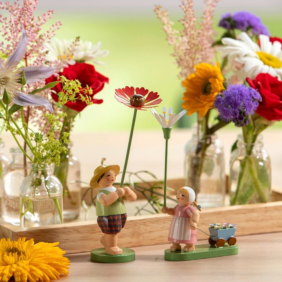 Zwei handgefertigte Holzfiguren von Blumenkindern auf einem Tisch vor mehreren Glasvasen mit bunten Blumen