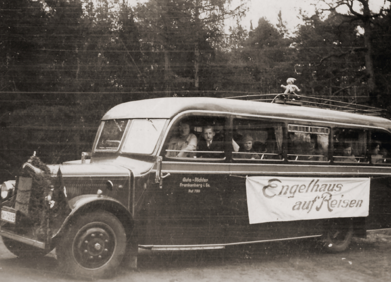 Schwarz -Weiß-Foto von historischem Reisebus mit einem Banner "Engelhaus auf Reisen". Auf dem Dach des Busses ist eine Wendt & Kühn-Figur angebracht.
