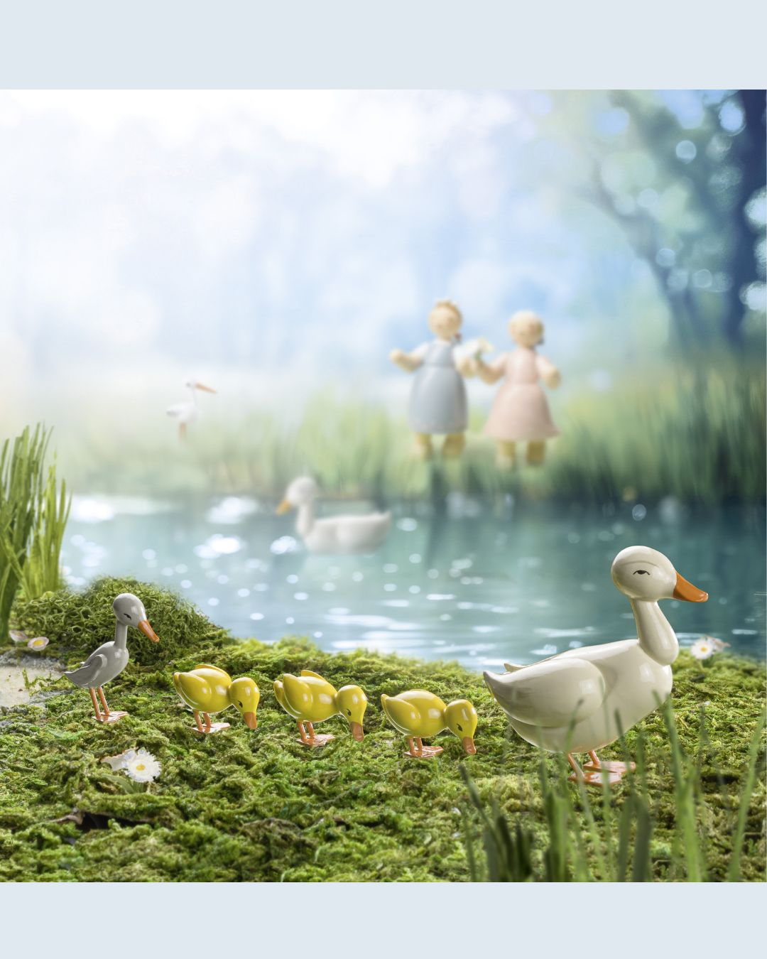 Handgefertigte Märchenfiguren von Wendt & Kühn zeigen eine Entenfamilie mit einem großen weißen Entlein und mehreren gelben Küken auf moosbedecktem Boden vor einem Teich, im Hintergrund zwei unscharfe Figuren von Kindern