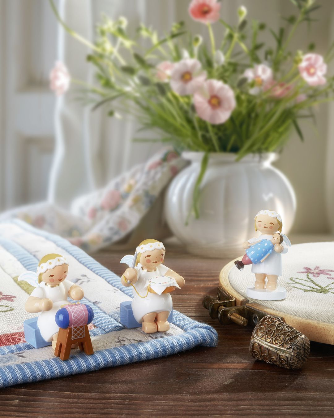 Drei handgefertigte Engel-Figuren von Wendt & Kühn bei Handarbeiten, dekoriert auf bestickten Stoffen und einer Holzoberfläche – liebevolle Geschenkidee für Handarbeitsfans, gefertigt in der Manufaktur in Grünhainichen im Erzgebirge
