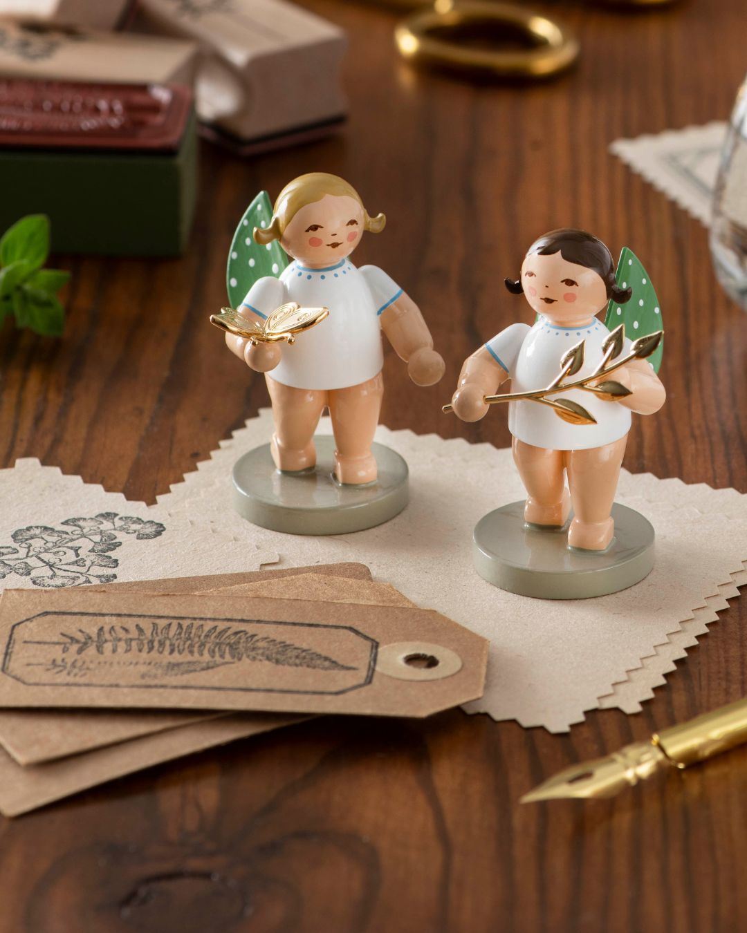 Zwei handgefertigte Engelfiguren aus Holz mit grünen Flügeln und weißen Kleidern, eine hält einen goldenen Schmetterling, die andere einen goldenen Zweig, auf grauen Sockeln stehend