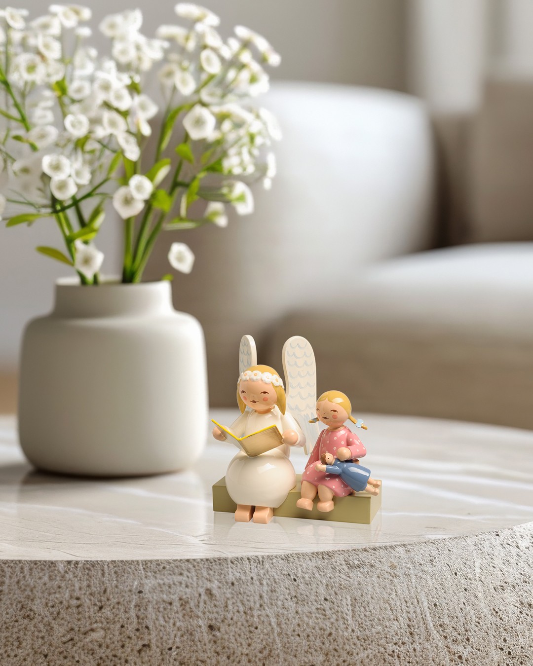 Handgefertigter Margeritenengel von Wendt & Kühn mit Mädchen auf einer Bank. Die Holzfigur aus Grünhainichen im Erzgebirge steht als hochwertige Wohnraumdekoration auf einem modernen Couchtisch.