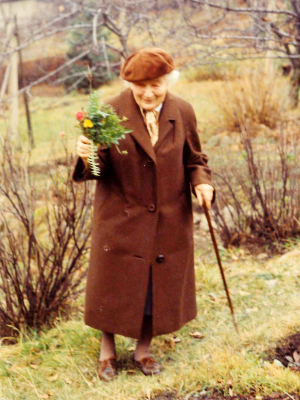 Grete Wendt im braunen Mantel und mit Mütze mit einem bunten Blumenstrauß in der Hand. Sie steht in einem sonst herbstlichen Garten.