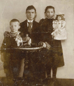 Schwarz-Weiß-Aufnahme. Grete im dunklen Kleid und mit Puppe im Arm neben ihren Brüdern. Curt im Anzug, Johannes als kleiner Junge im Ringelpullover.