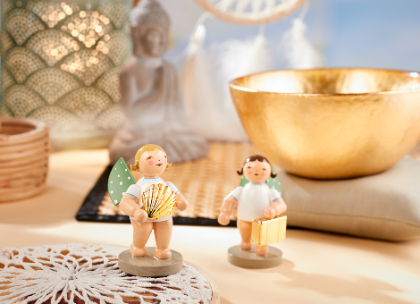 Figuren "Weltenbummler" mit Koffer und "Entdecker" mit Muschel in einer Boho-Dekoration. Im Hintergrund stehen eine goldene Klangschale und Teelichter. Außerdem liegen Makramee-Stücke aus.