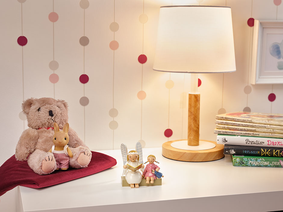 Margeritenengel auf Bank mit Mädchen auf einer weißen Kinderzimmerkommode. Darauf stehen außerdem Kuscheltiere, eine Lampe und Bücher. Die Tapete des Zimmers ist in Rosatönen gepunktet.