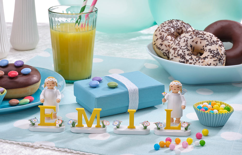 Zwei Margeritenengel flankieren den Namen „Emil“. Das Ensemble steht auf einer Geburtstagstafel mit Limonade, Doughnuts und Geschenken. Das Geschirr und die Tischdecke ist blau.