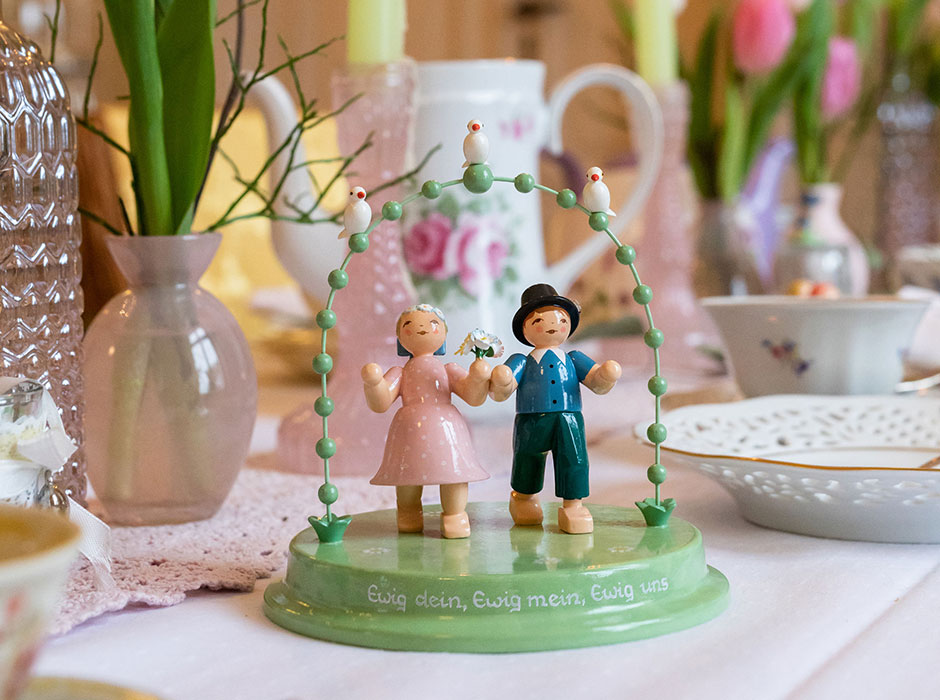 Liebespaar (Junge und Mädchen) im Bogen auf einer gedeckten Tafel mit Tulpen-Gestecken. Das Geschirr ist mit rosafarbenen Rosen gemustert.