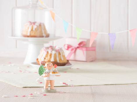 Engel mit Kuchen und Herz auf einem weißen Holztisch. Im Hintergrund stehen zwei Gugelhupf-Kuchen. Einer davon unter einer Glocke. Daneben liegt ein in rosafarbenes Papier verpacktes Geschenk.
