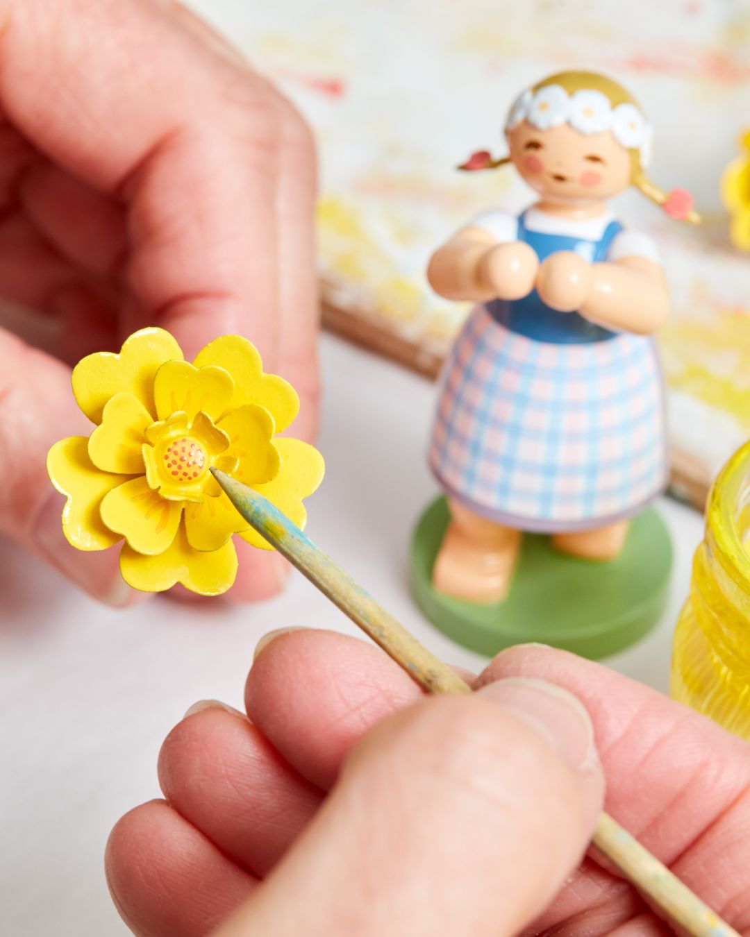 Hand, die Blume aus Holz in gelber Farbe bemalt. Im Hintergrund eine Figur.