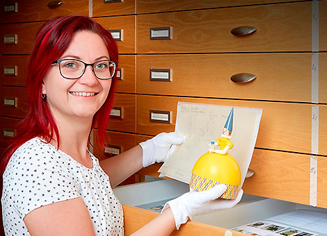Die Kuratorin Marlis Rokitta vor dem Archivschrank. Sie zeigt ein Dokument und die Figur "Knauldame" im gelben Kleid.