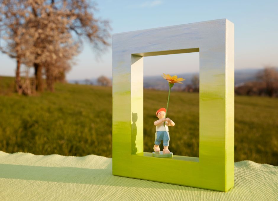 Grün und Hellblau bemalter Holzrahmen mit Figur mit Blüte vor Obstwiese