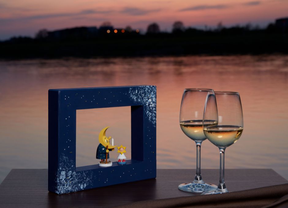 Blauer Holzrahmen mit zwei Figuren, ein Mondvater und ein Sternenkind, vor Abendszene am See