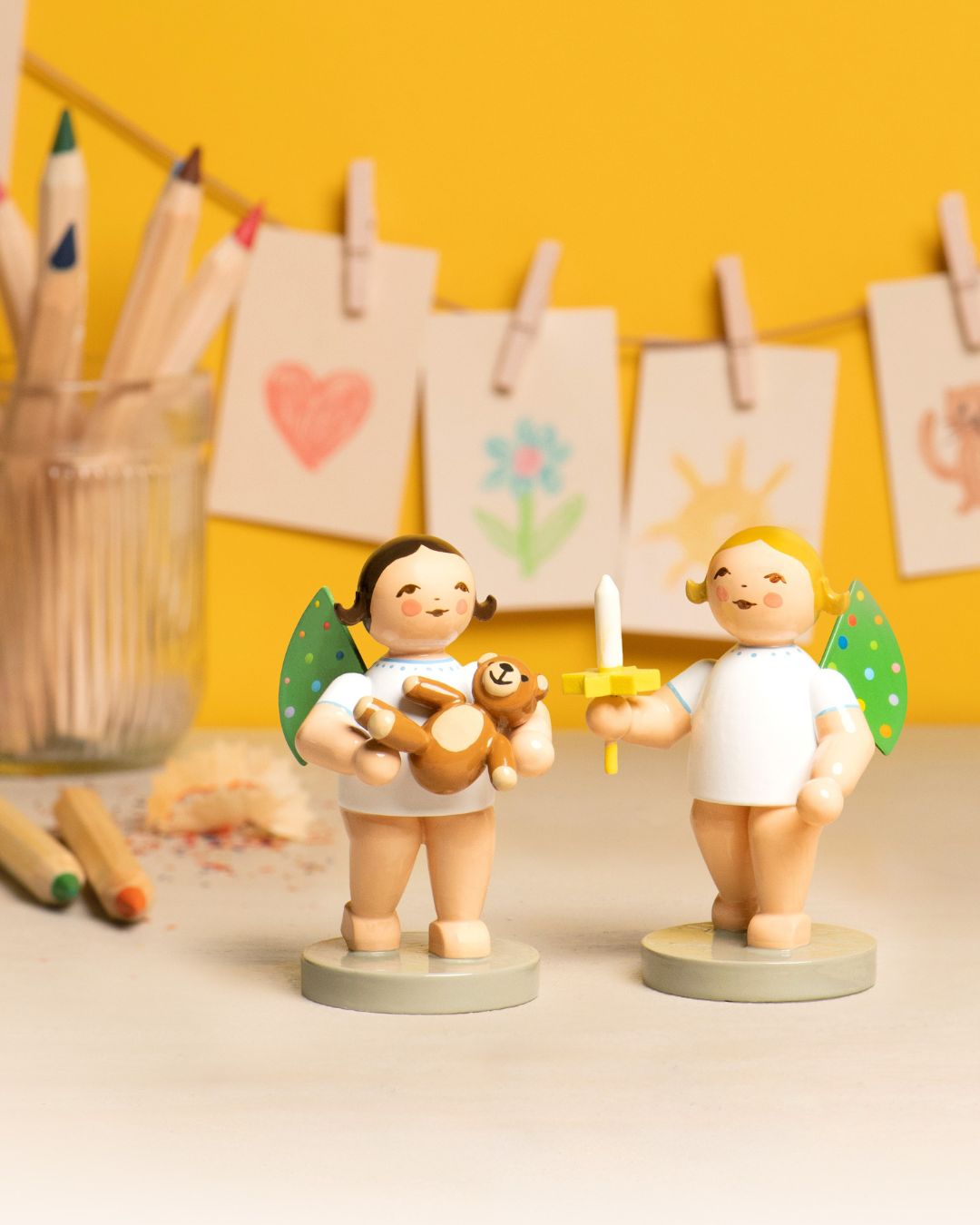 Zwei handgefertigte Engel aus Holz mit grünen Flügeln, einer hält einen Teddybär, der andere eine Kerze, vor gelbem Hintergrund mit an einer Leine hängenden Kinderzeichnungen