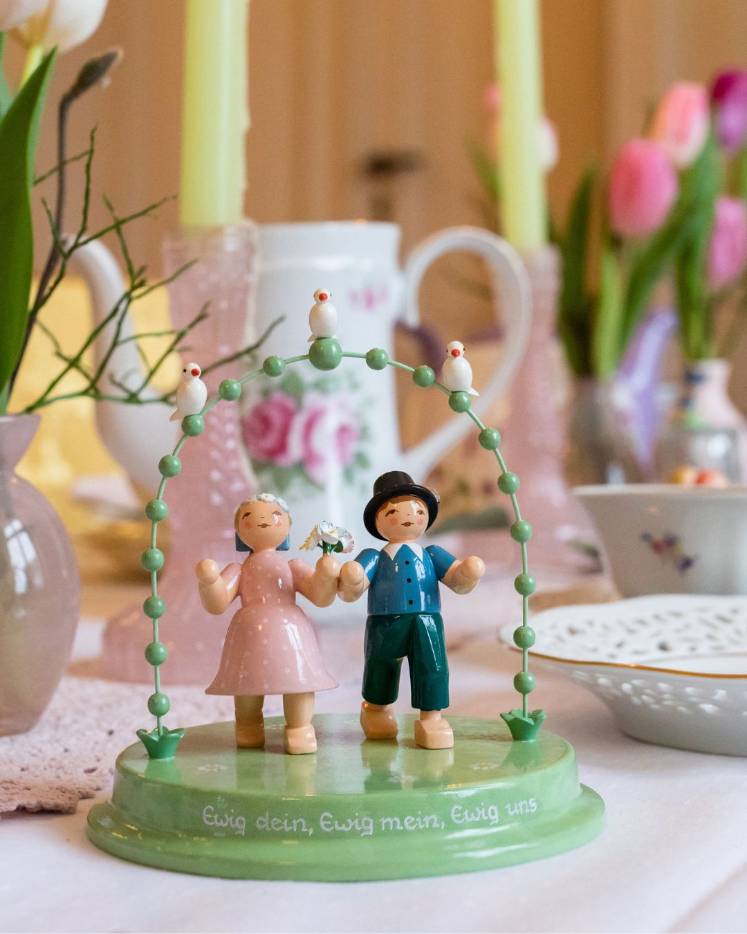 Zwei Holzfiguren eines Liebespaars unter einem grünen Bogen auf einem gedeckten Tisch mit Kerzen und Blumen im Hintergrund