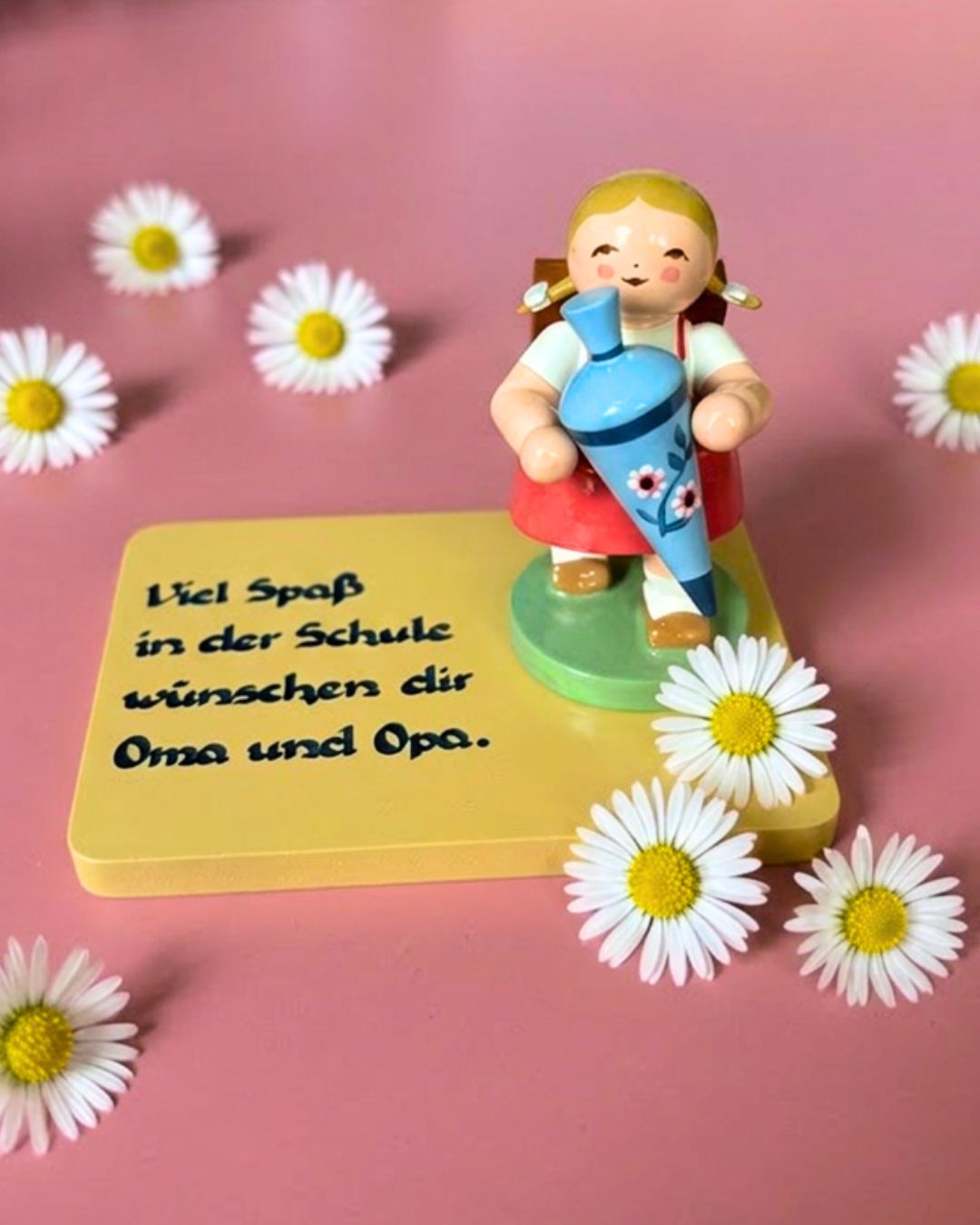 Figur eines Mädchens mit Zöpfen und rotem Kleid, das eine blaue Schultüte hält, auf gelbem Sockel mit der schwarzen Schrift 'Viel Spaß in der Schule wünschen dir Oma und Opa', umgeben von Gänseblümchen auf rosa Untergrund