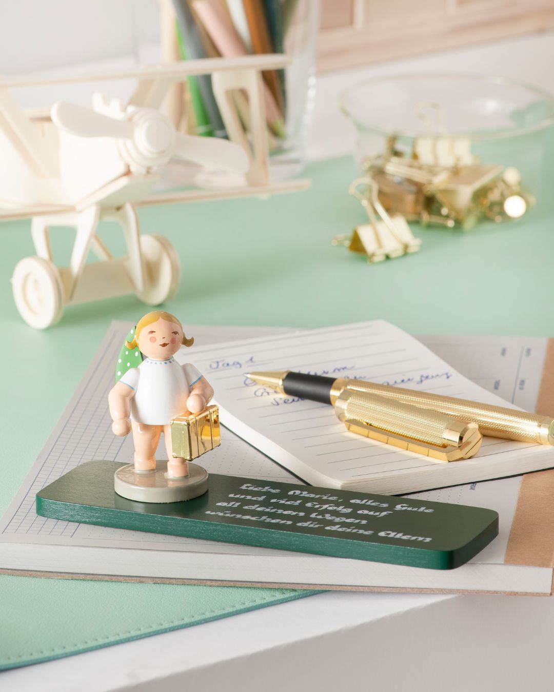 Handgefertigte Figur aus Holz eines kleinen reisenden Engels mit Koffer auf grüner Sockelplatte mit goldfarbenem Stift und Notizzettel im Hintergrund